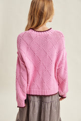 woman wearing a pink oversized knit sweater with a diamond pattern, shown from the back over a plaid skirt