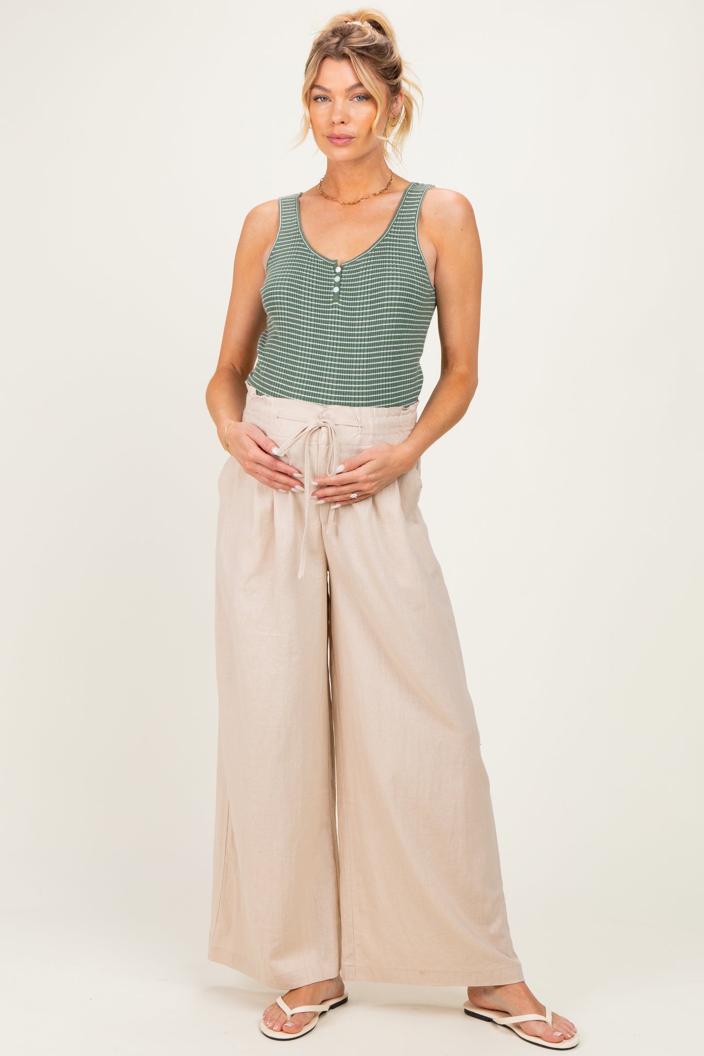 woman wearing a green plaid maternity tank top and beige wide-leg pants, holding her baby bump