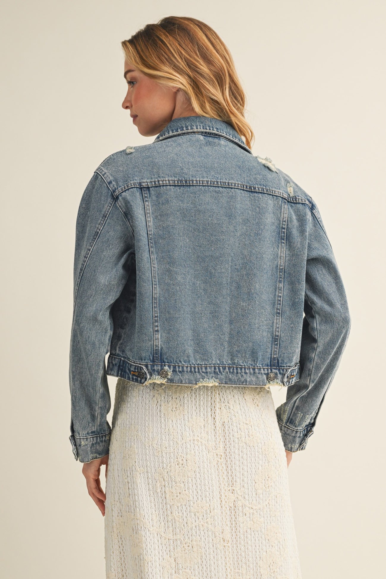 woman wearing a distressed denim jacket with a cropped cut, styled over a lace dress, showing the back view