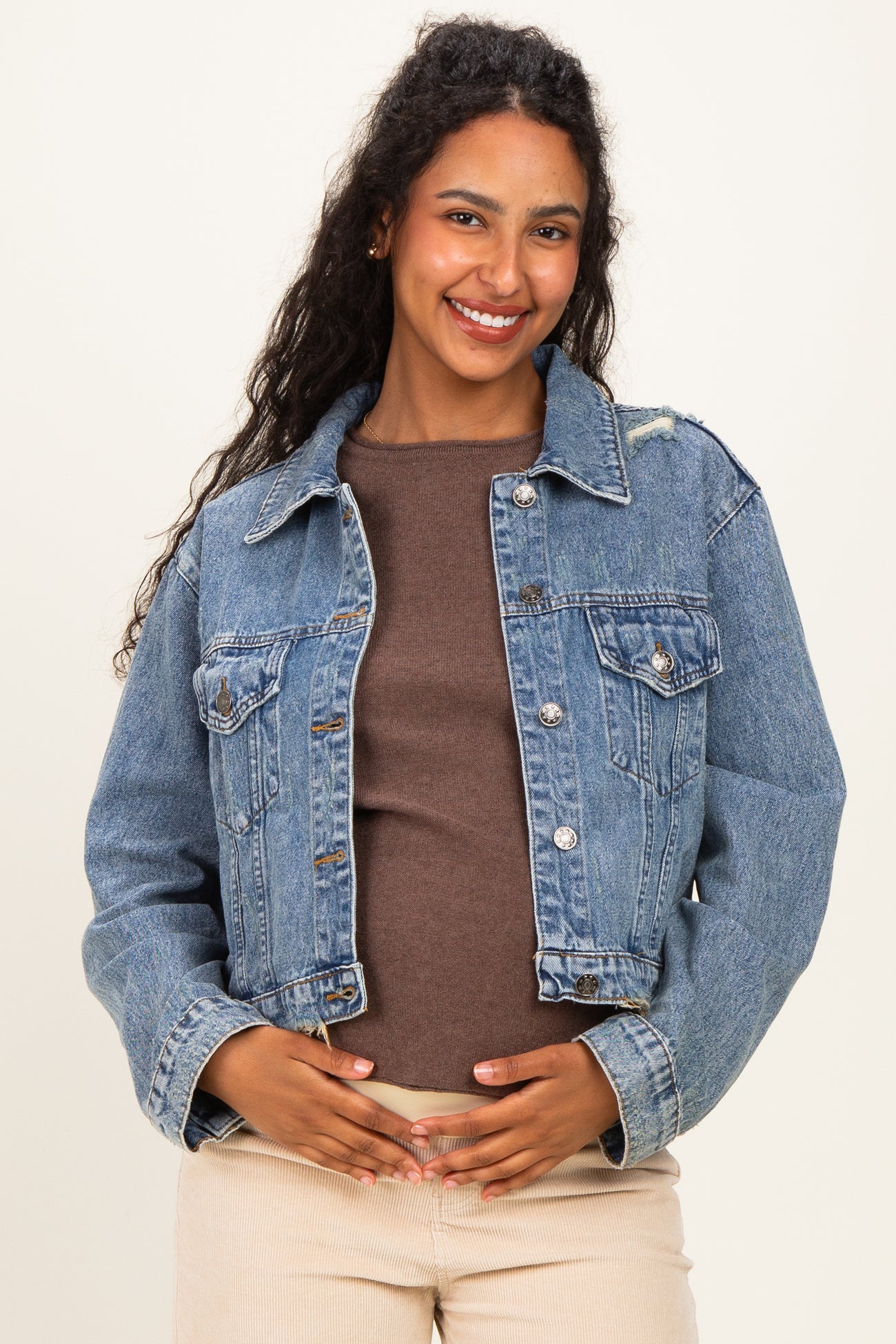 woman wearing a light blue maternity denim jacket over a brown top, gently holding her baby bump