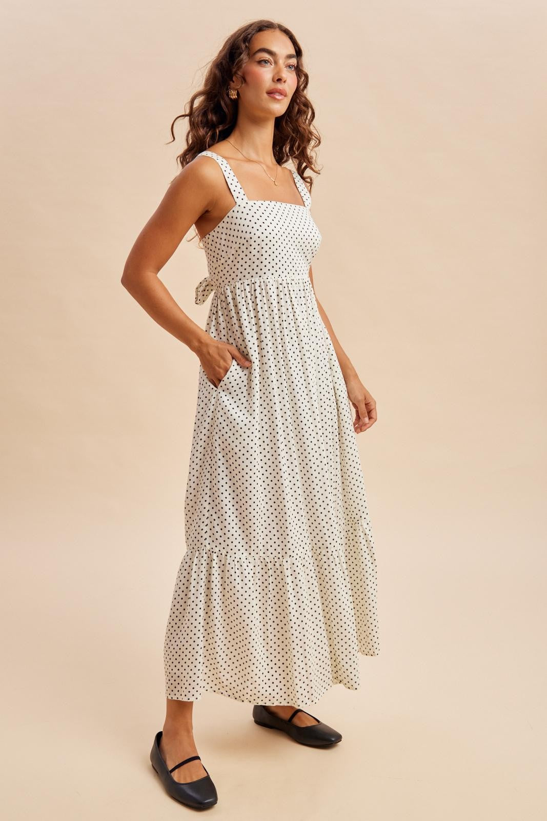 woman wearing a white polka dot maxi dress with square neckline and tie back detail, standing in a relaxed pose