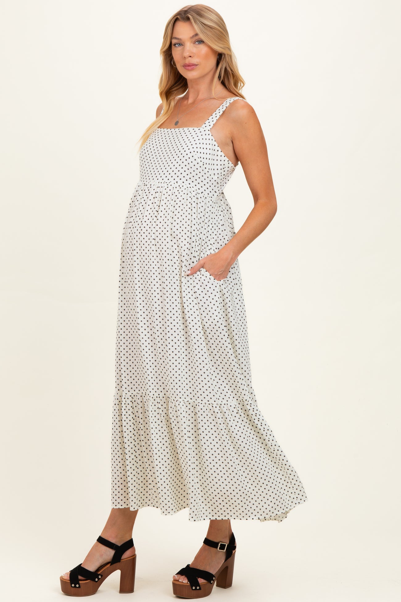 woman wearing a white polka dot maternity dress with a tiered skirt and pockets, standing in platform sandals