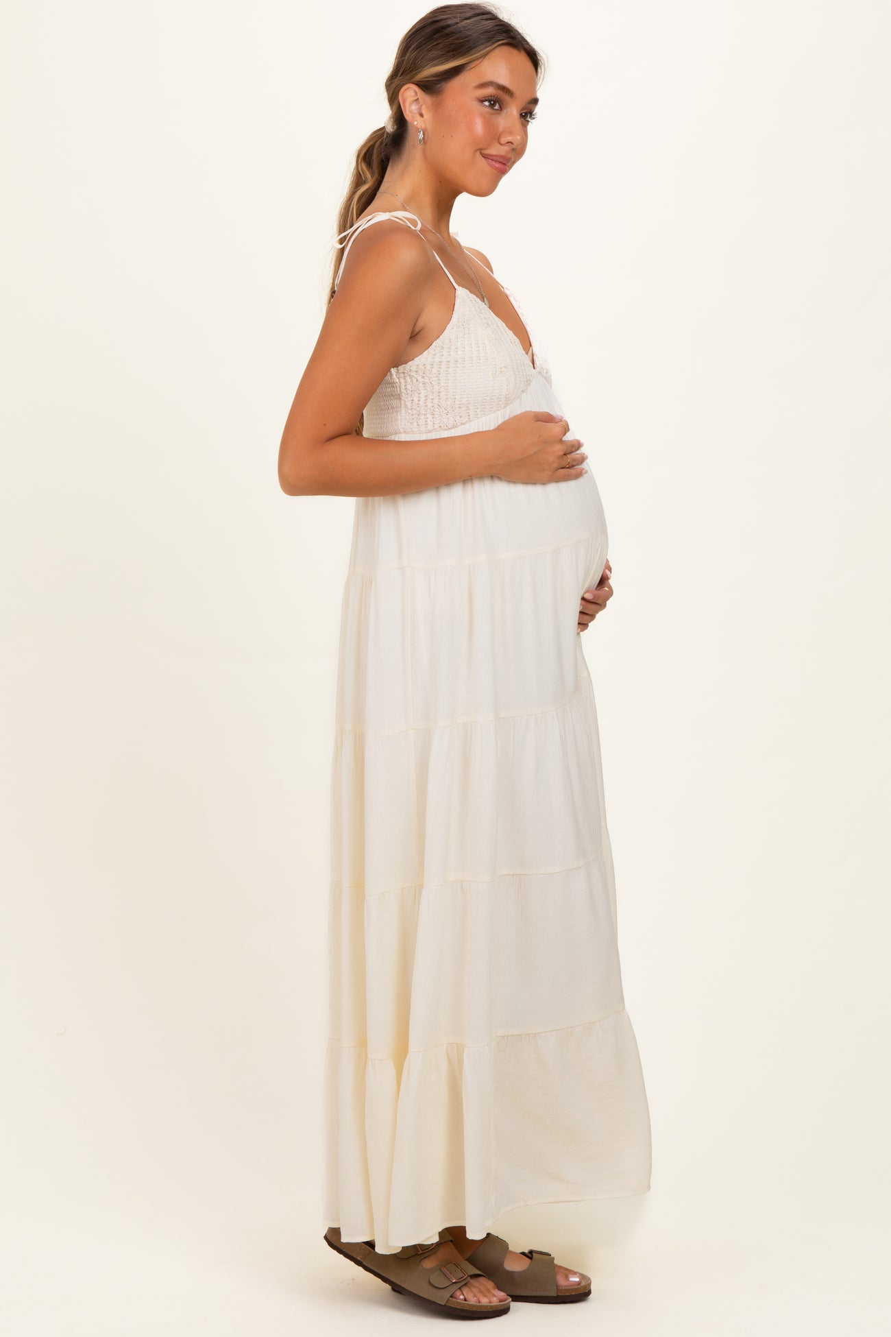 woman wearing a cream tiered maternity maxi dress with a fitted bodice, holding her baby bump