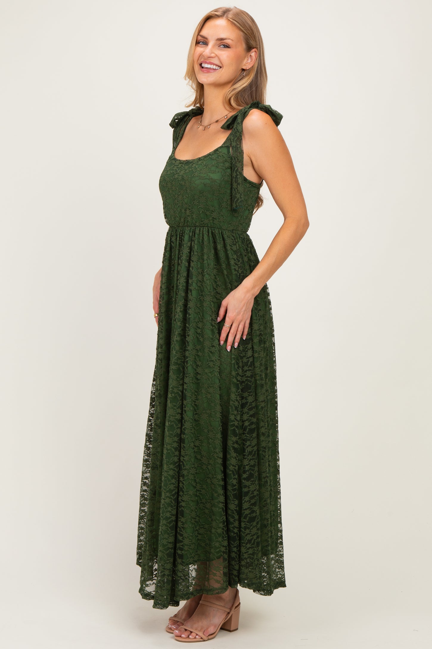 woman wearing a dark green lace maxi dress with tie straps and a fitted waist, smiling at the camera