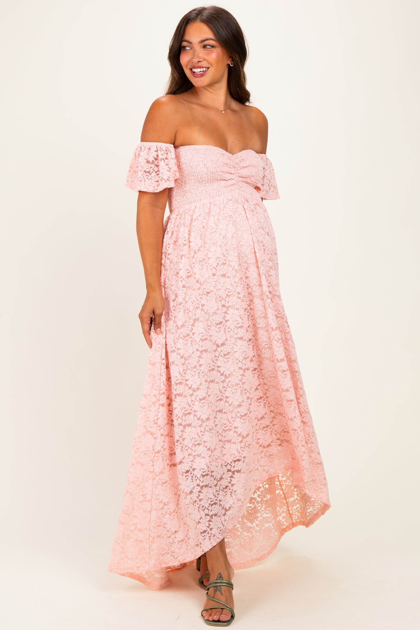 woman wearing a pink off-the-shoulder lace maternity dress with a fitted bodice and flowy skirt, showcasing her baby bump