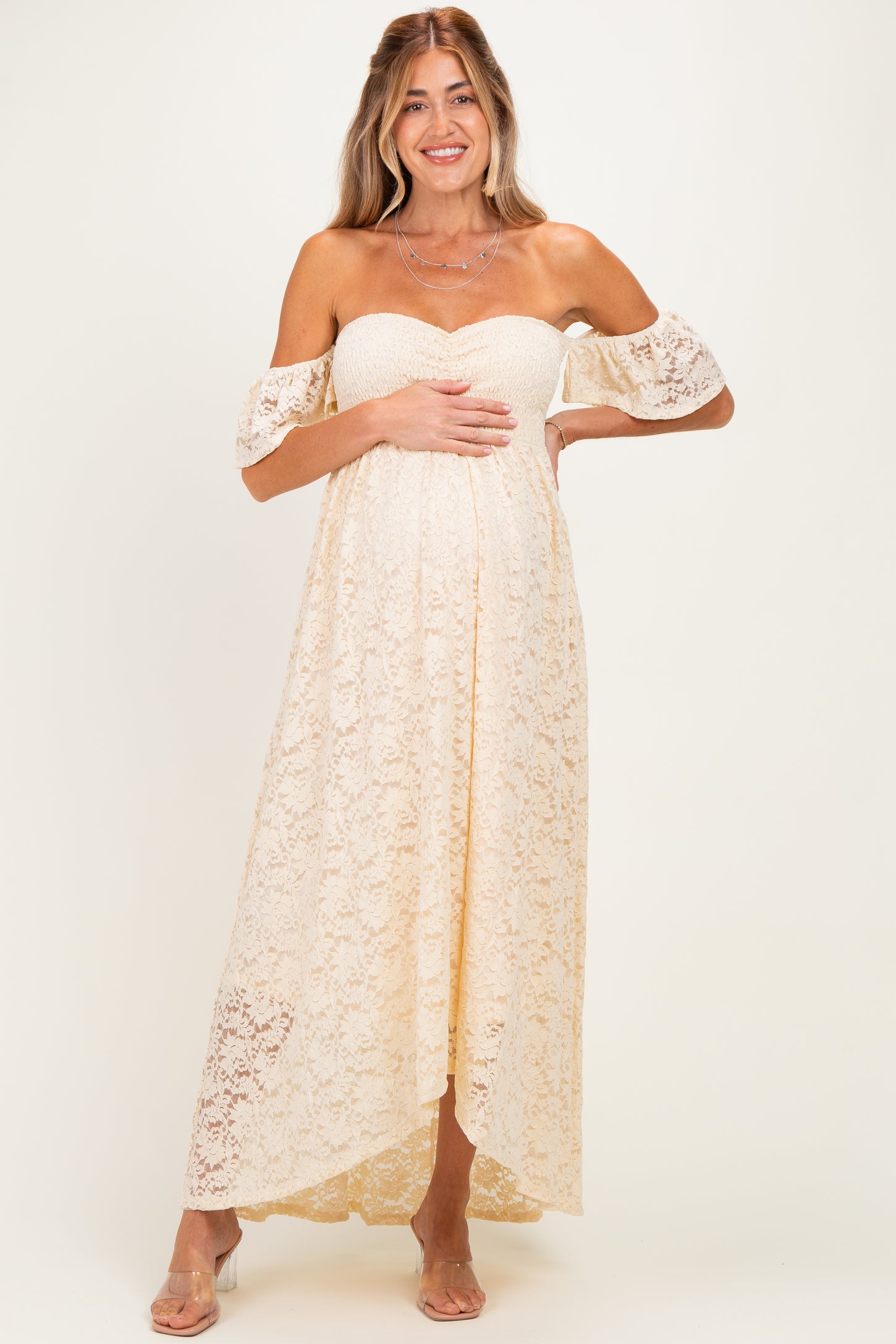 woman wearing a cream lace off-shoulder maternity dress and holding her baby bump