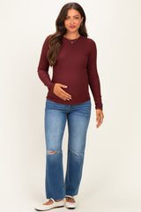 woman wearing a burgundy long sleeve maternity top and distressed blue jeans, gently cradling her baby bump