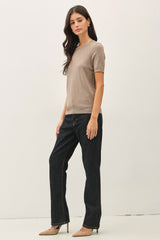 woman wearing a beige short sleeve knit top and dark denim straight leg jeans, standing in a neutral background