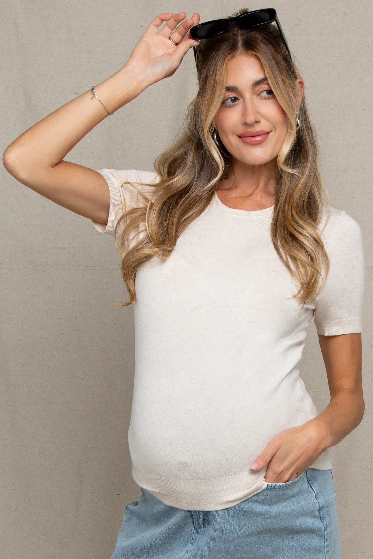 woman wearing a light beige short sleeve maternity top and blue skirt, showing her baby bump and holding sunglasses