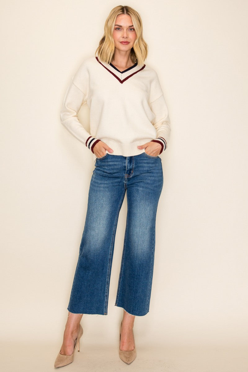 woman wearing a white v-neck sweater with burgundy stripes and blue wide-leg jeans, standing in a neutral background
