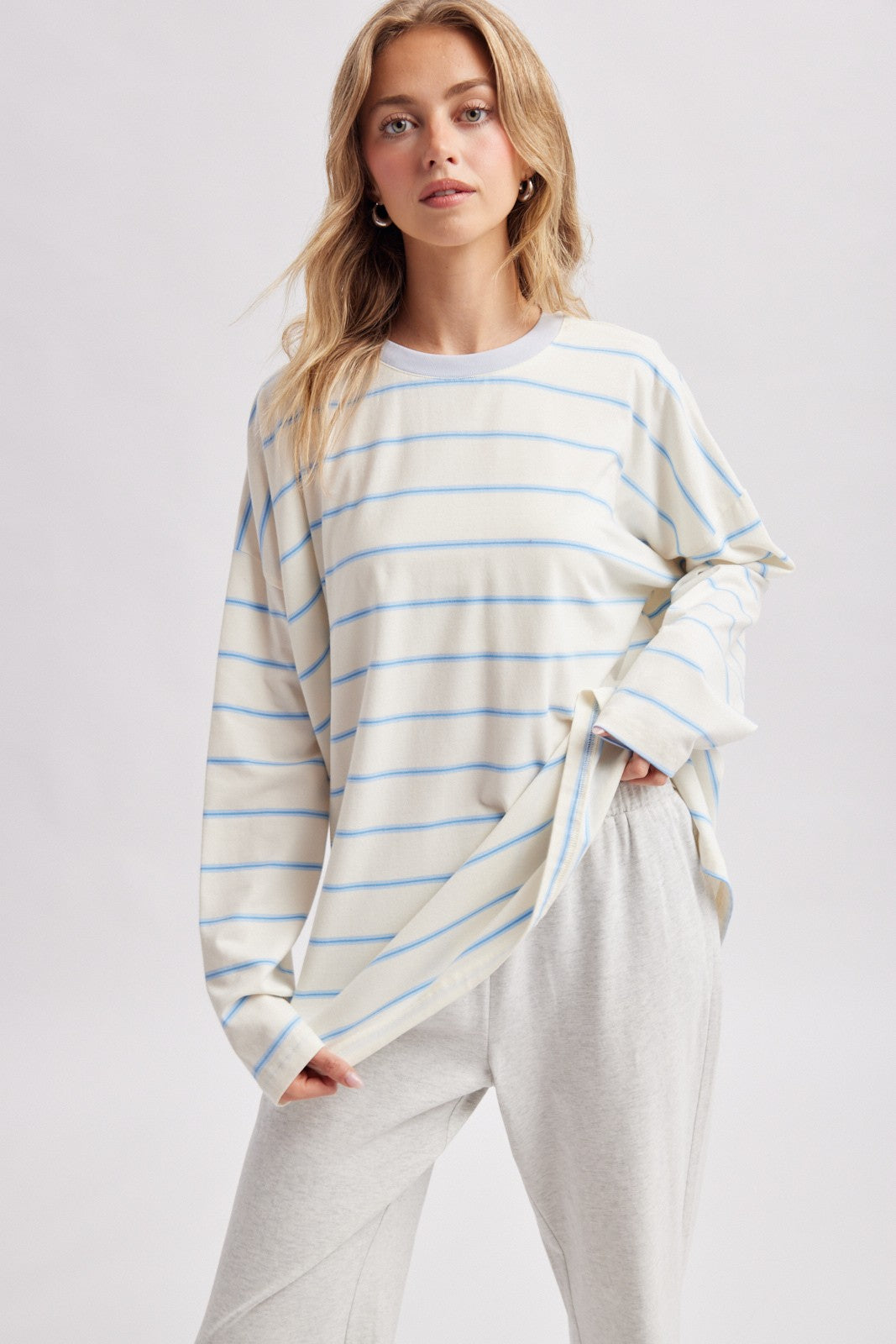 woman wearing a cream and light blue striped long sleeve top with relaxed fit and holding the hem, paired with gray pants