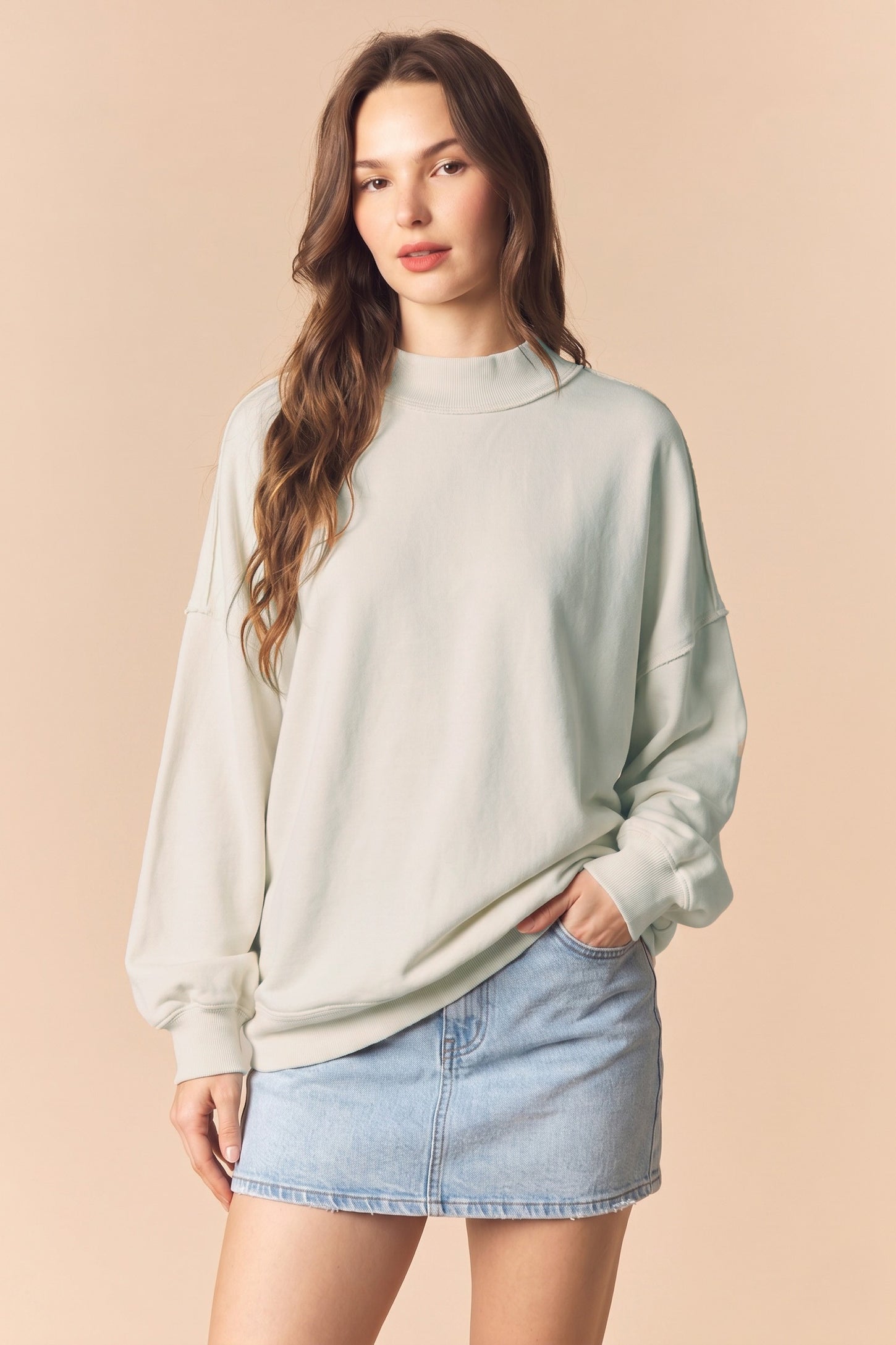 woman wearing a light green oversized sweatshirt and a denim mini skirt, hands in pockets, with a neutral background