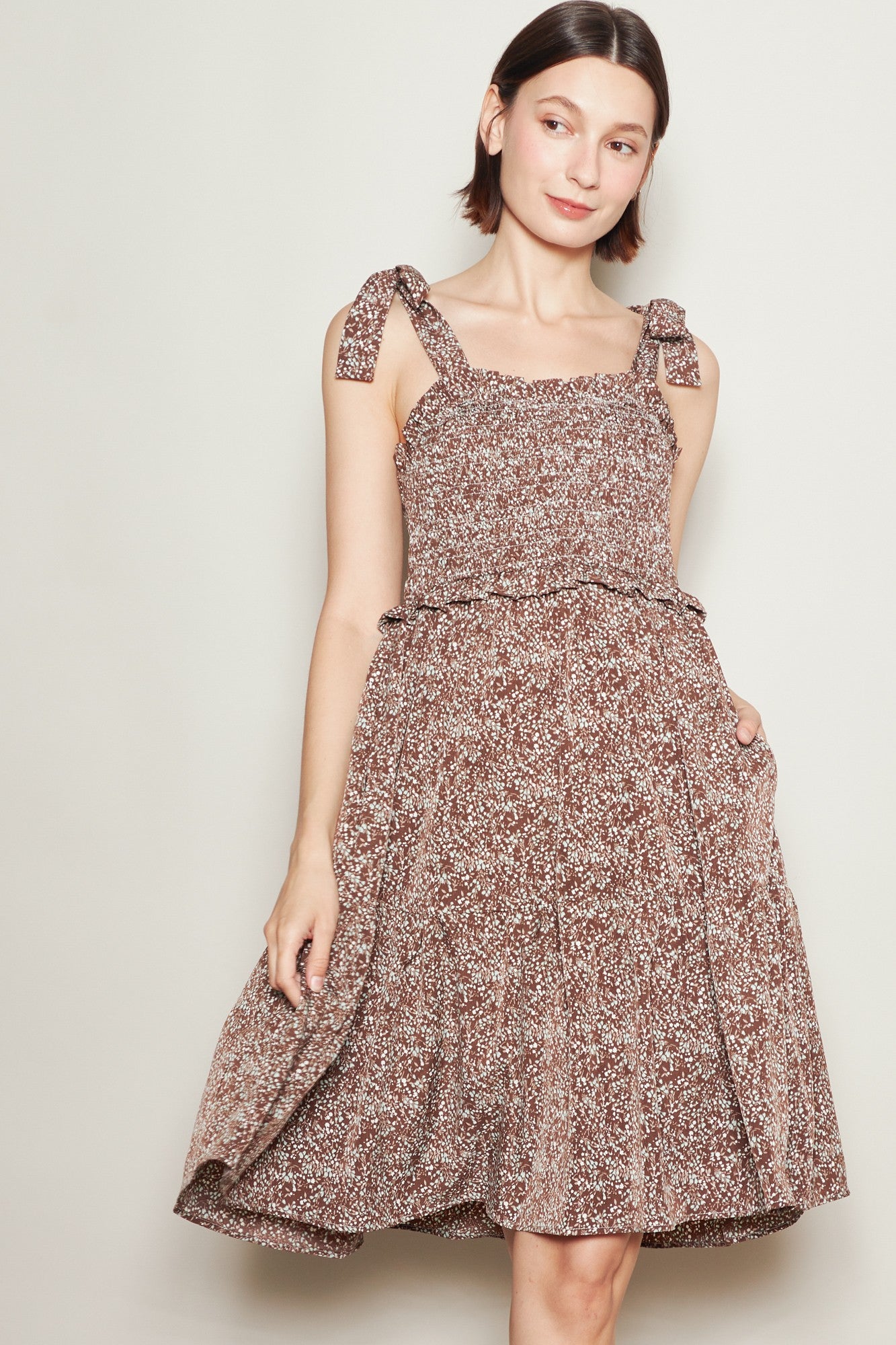 woman wearing a brown floral sundress with tie straps and a flared skirt, standing with one hand in her pocket