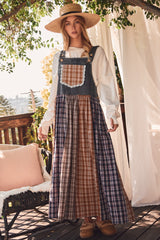 woman wearing a long plaid overall dress with denim top and tiered skirt, standing on a porch among greenery