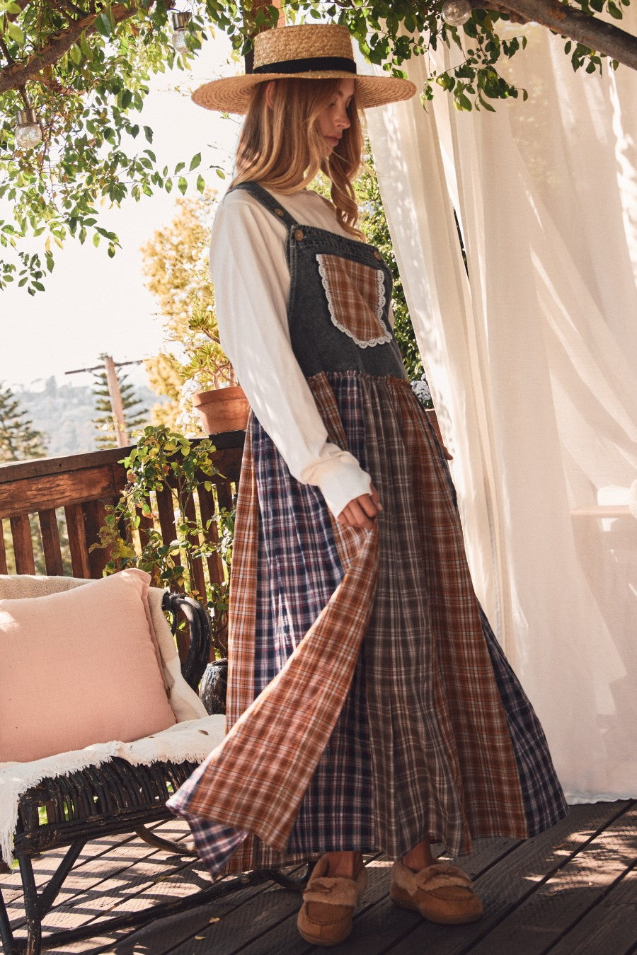 woman wearing a denim overalls dress with a plaid skirt and a white long sleeve top, standing outdoors in natural light
