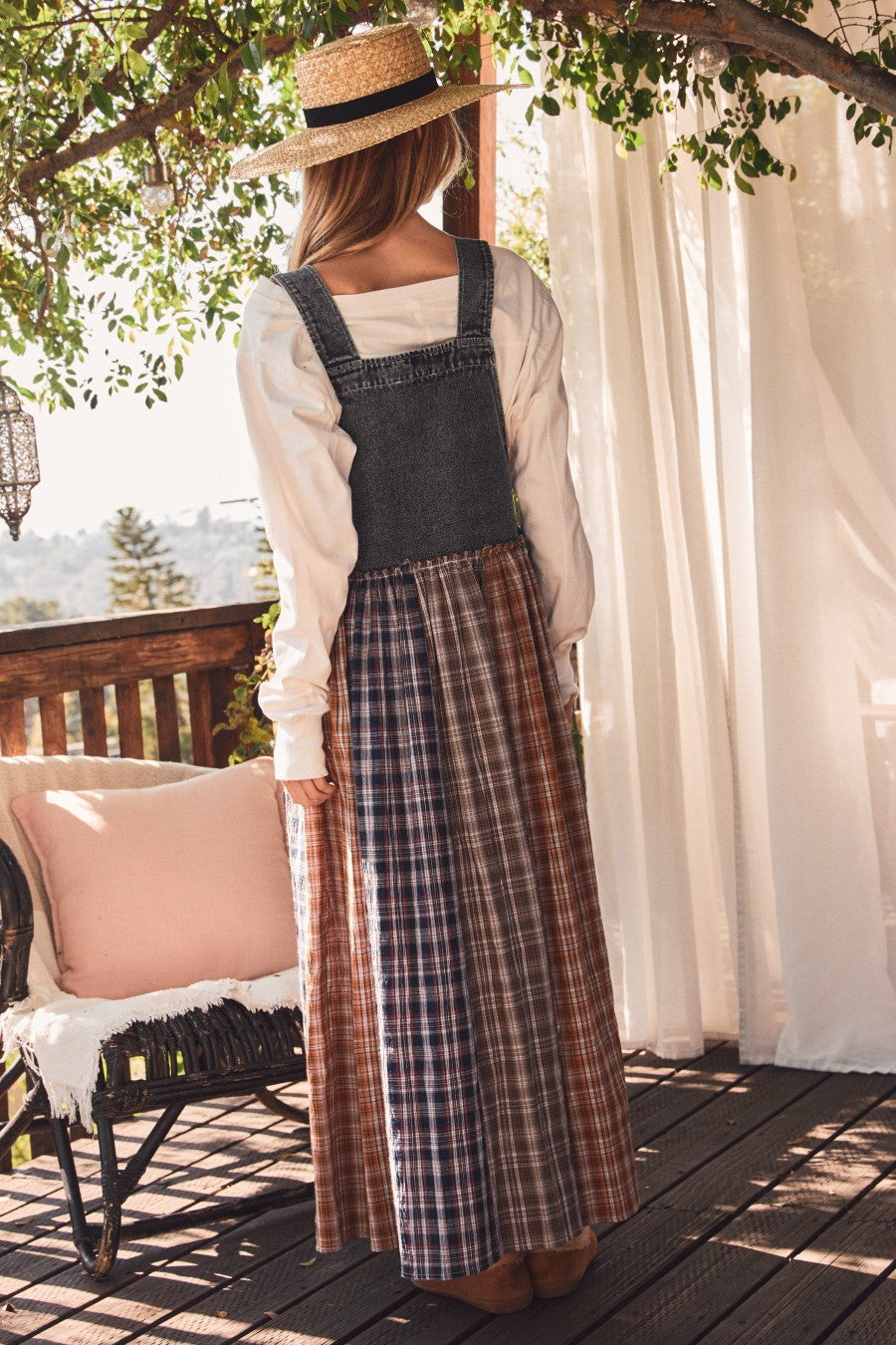 woman wearing a denim bib dress with a plaid maxi skirt, standing on a deck with a straw hat and a sunlit background