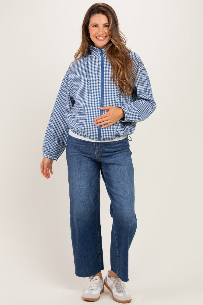 woman wearing a blue and white gingham maternity jacket and blue wide-leg jeans, gently cradling her baby bump
