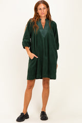 woman wearing a dark green corduroy tunic dress with a v-neck and pockets, styled with black slip-on shoes