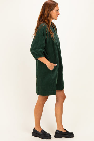 woman wearing a dark green corduroy dress with three-quarter sleeves and pockets, standing sideways