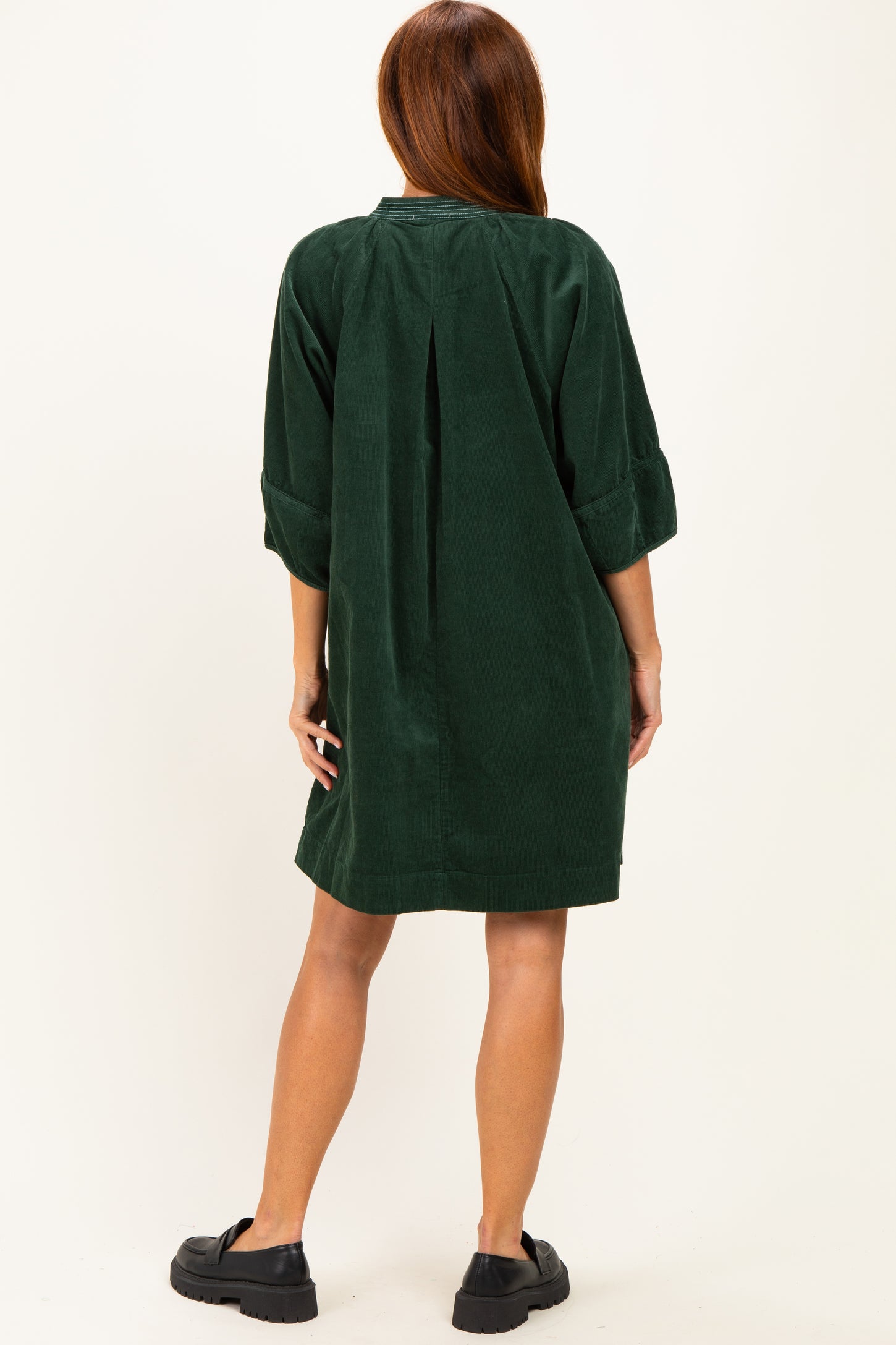 woman wearing a dark green corduroy shirt dress with three-quarter sleeves and a back pleat, styled with black clogs