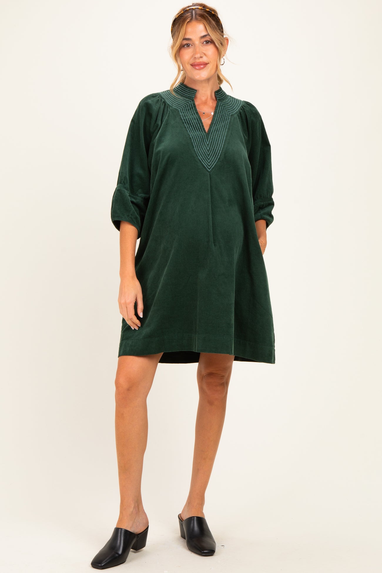 woman wearing a dark green maternity dress with decorative stitching and puff sleeves, standing against a light background