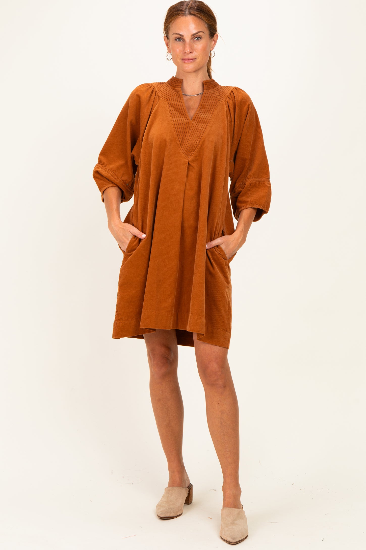 woman wearing an oversized rust-colored dress with pleated details and side pockets, standing against a neutral background