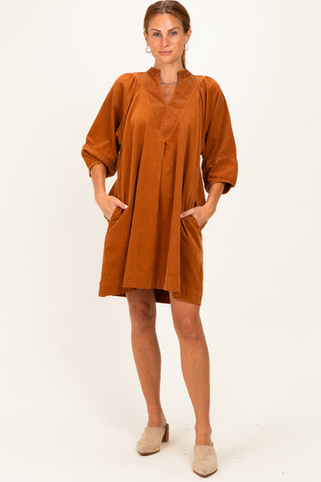 woman wearing an oversized rust-colored dress with pleated details and side pockets, standing against a neutral background