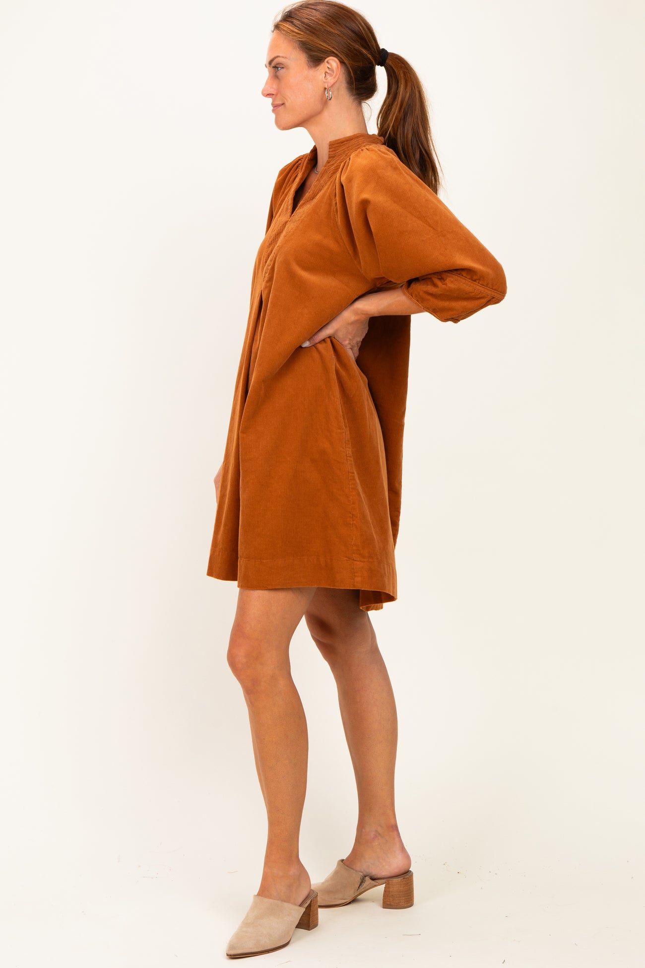 woman wearing a rust-colored corduroy shirt dress with a relaxed fit and three-quarter sleeves, styled with beige mules