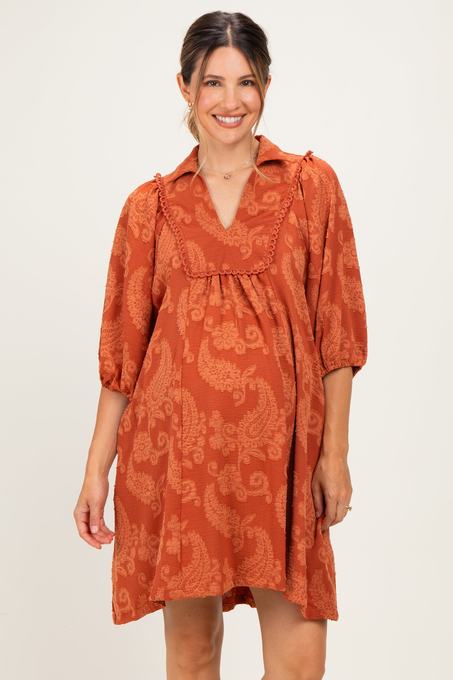 woman wearing a rust-colored maternity dress with a collar, puff sleeves, and paisley print, showing her baby bump