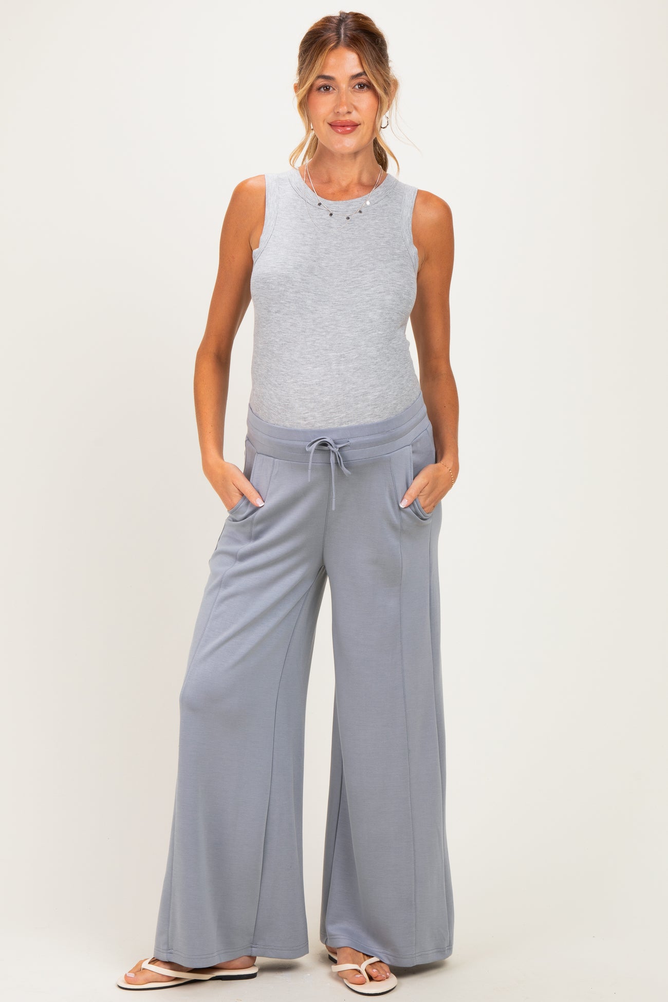 woman wearing a light gray sleeveless maternity top and wide-leg maternity pants with pockets, showing her baby bump