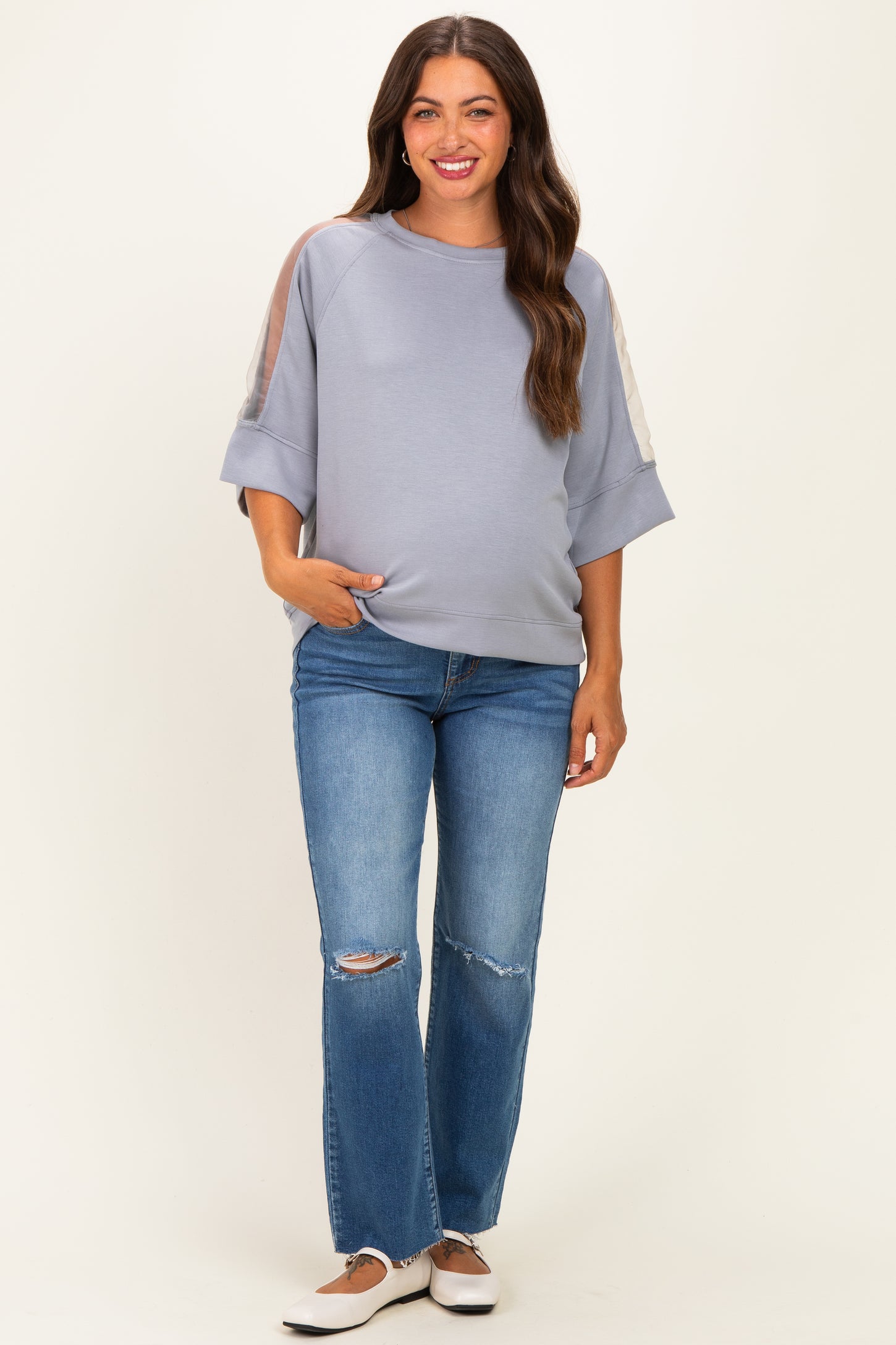 woman wearing a light blue maternity sweatshirt with sheer sleeves and pocket, paired with distressed jeans
