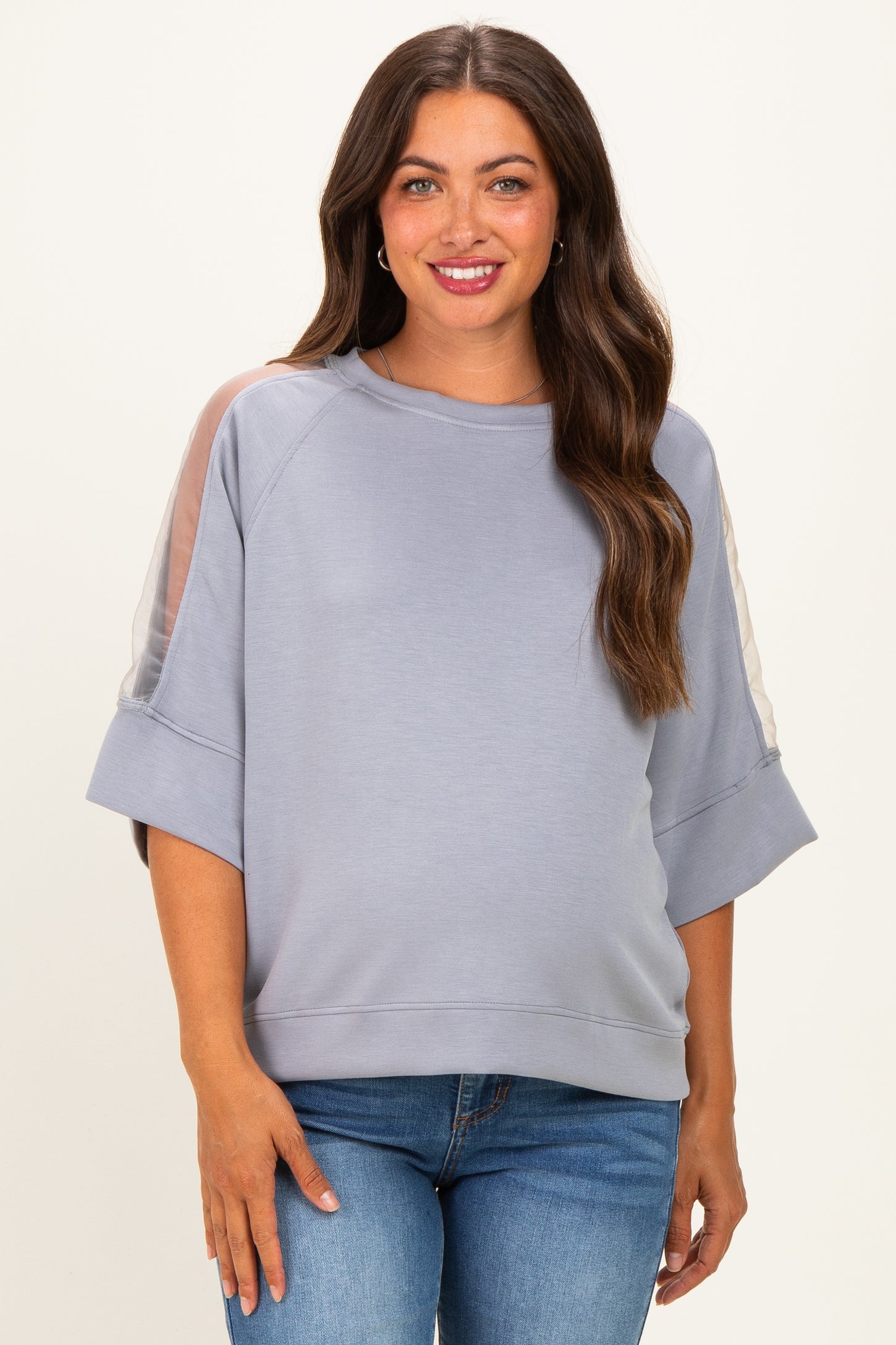 woman wearing a light grey maternity sweatshirt with sheer panels on the sleeves and a relaxed fit, smiling at the camera