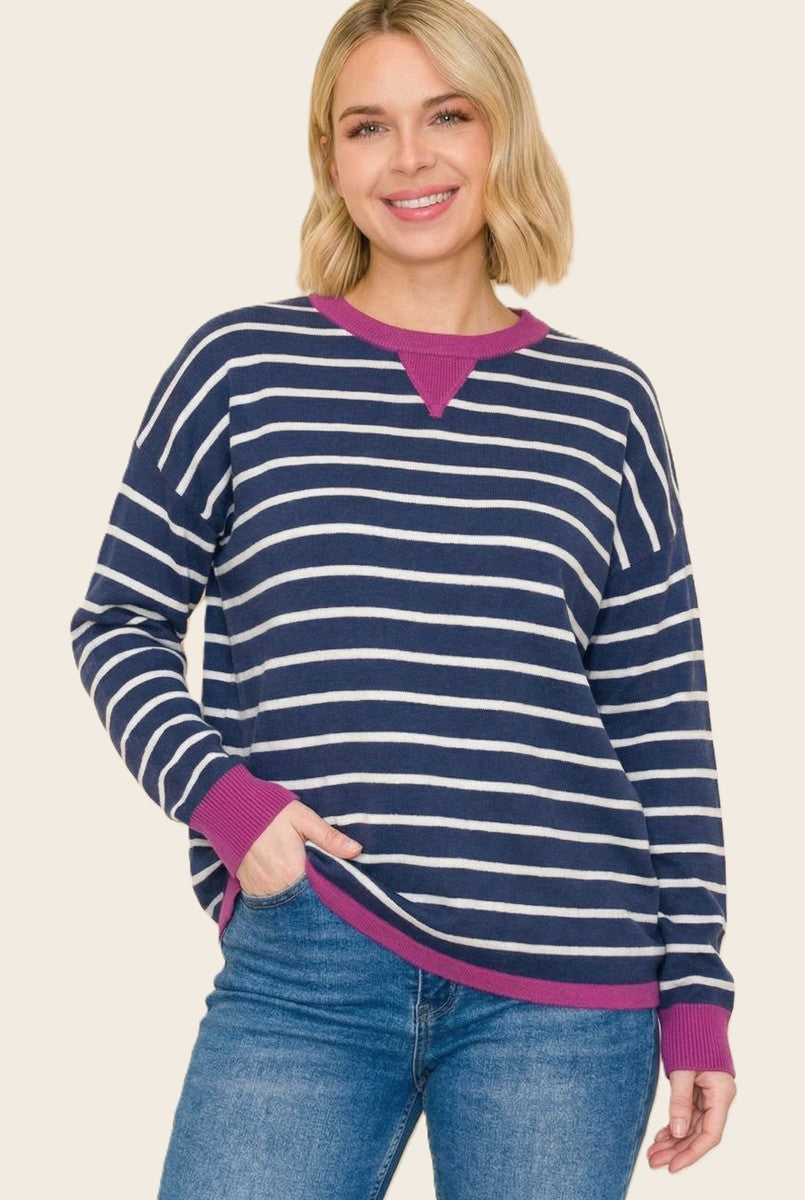 woman wearing a navy blue and white striped long sleeve top with a pink neckline and cuffs, smiling while standing with one hand in her pocket