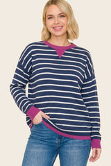 woman wearing a navy blue and white striped long sleeve top with a pink neckline and cuffs, smiling while standing with one hand in her pocket