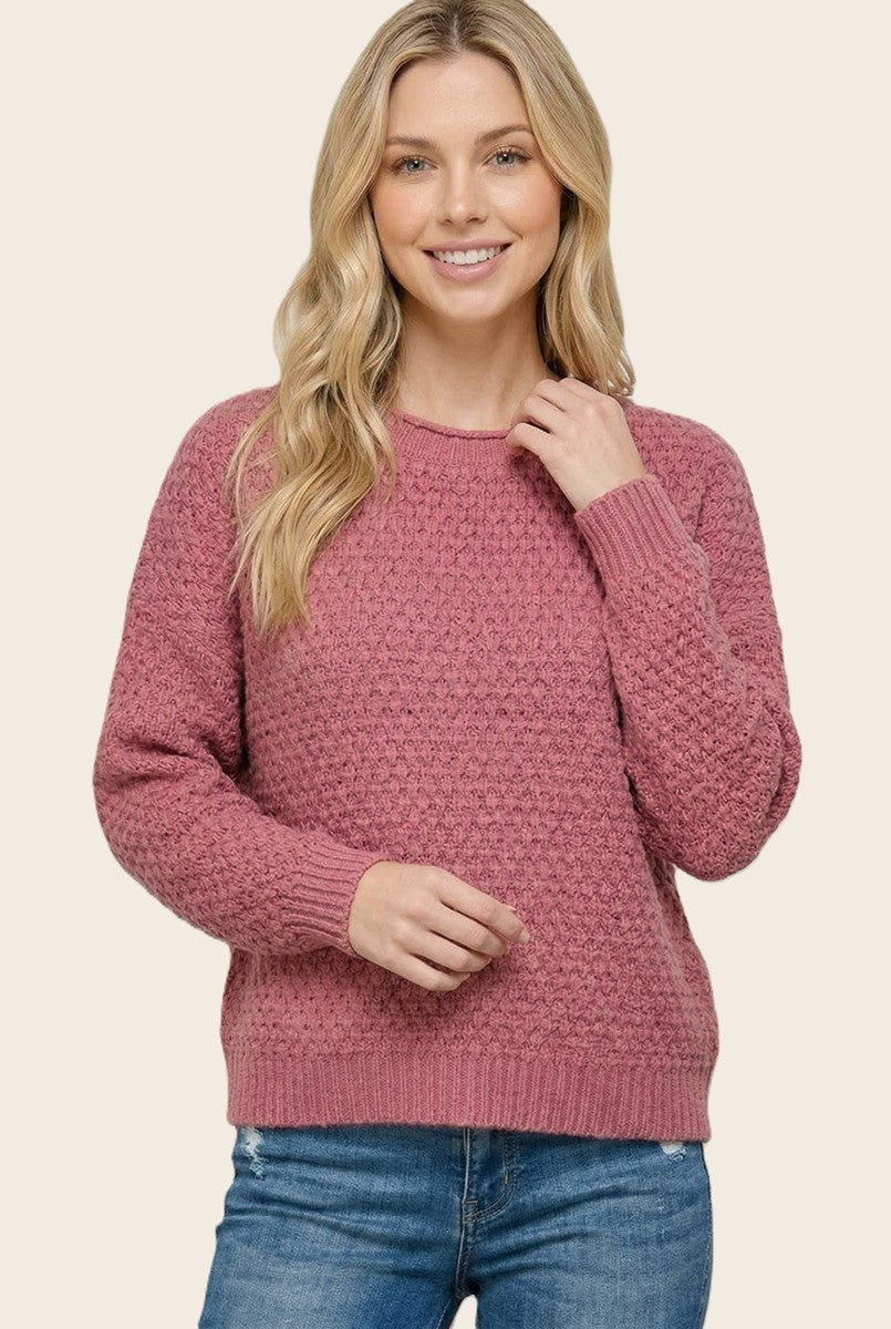 woman wearing a rose pink textured sweater and blue jeans, smiling and posing with one hand on her shoulder