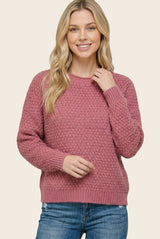 woman wearing a rose pink textured sweater and blue jeans, smiling and posing with one hand on her shoulder
