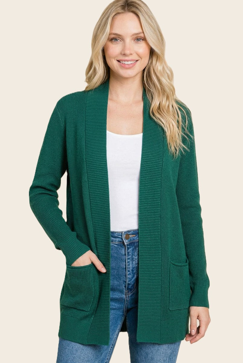 woman wearing a green long sleeve cardigan with pockets and a white top, smiling against a neutral background