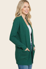 woman wearing a dark green open front cardigan with pockets and a white top, smiling against a neutral background