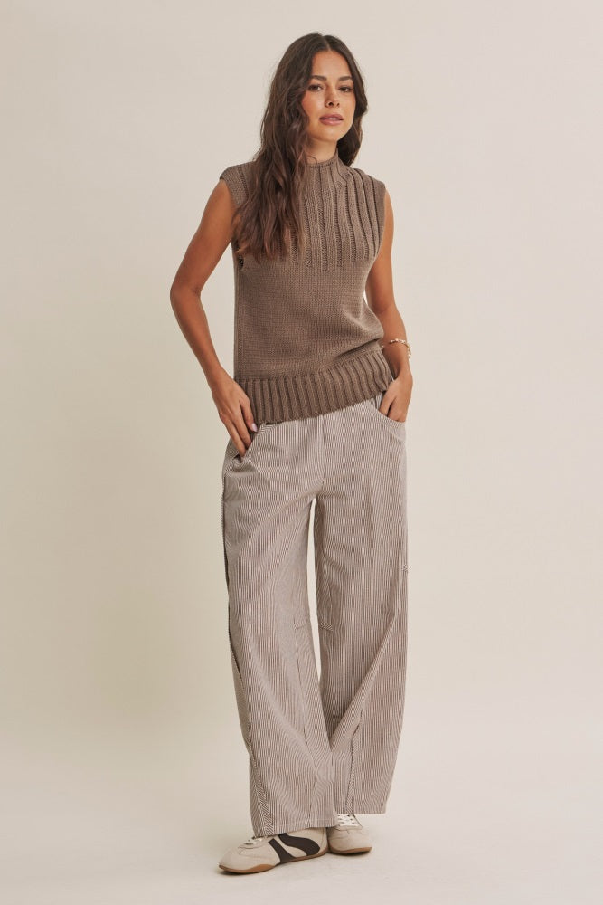 woman wearing a brown sleeveless textured sweater and striped wide-leg pants, standing with hands in pockets