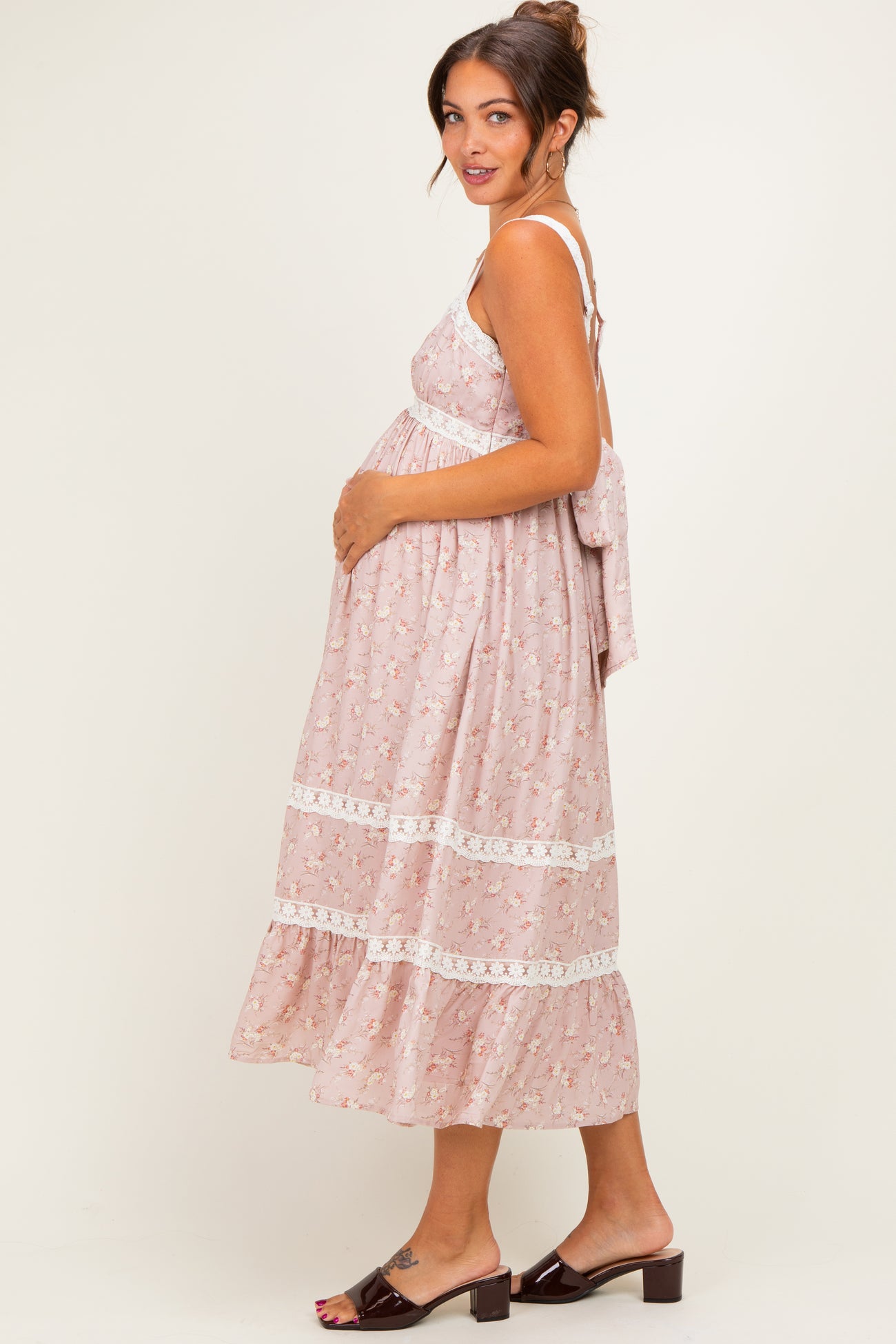 woman wearing a pink floral maternity dress with lace details and adjustable straps, holding her baby bump
