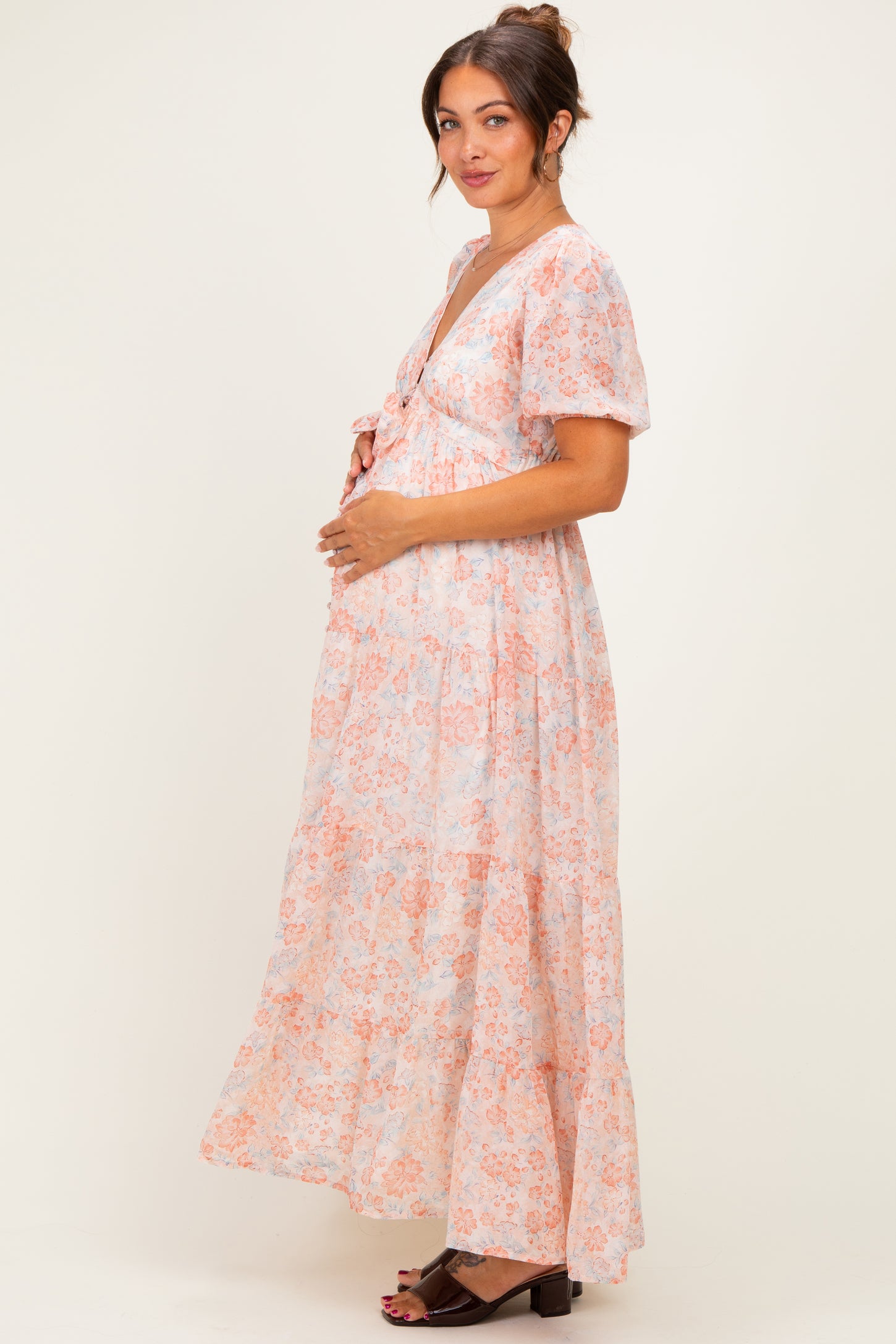 woman wearing a pink and orange floral maternity dress with a tie front, standing and holding her baby bump