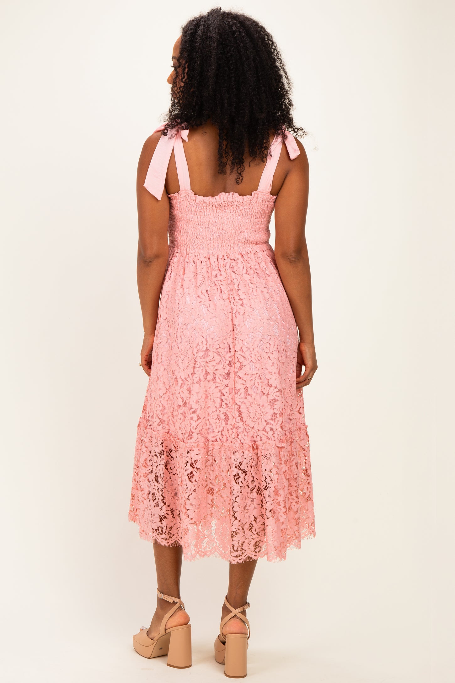 woman wearing a pink lace midi dress with smocked bodice and tie straps, standing with her back to the camera
