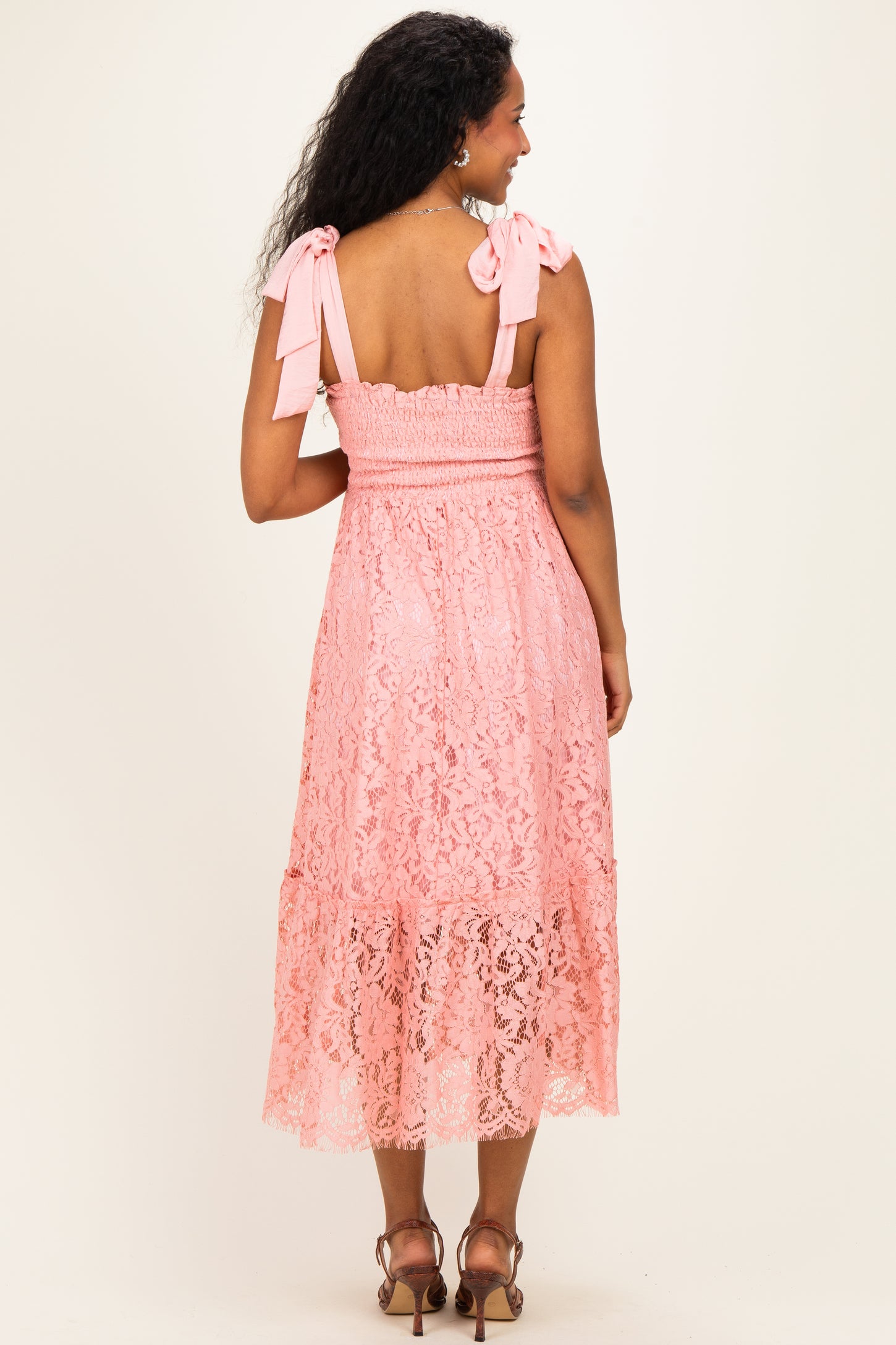 woman wearing a pink lace maternity dress with bow tie straps and a tiered hem, showing a baby bump from the back