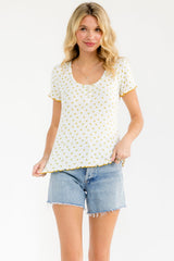 woman wearing a white short sleeve top with yellow floral print and scalloped trim, paired with denim shorts