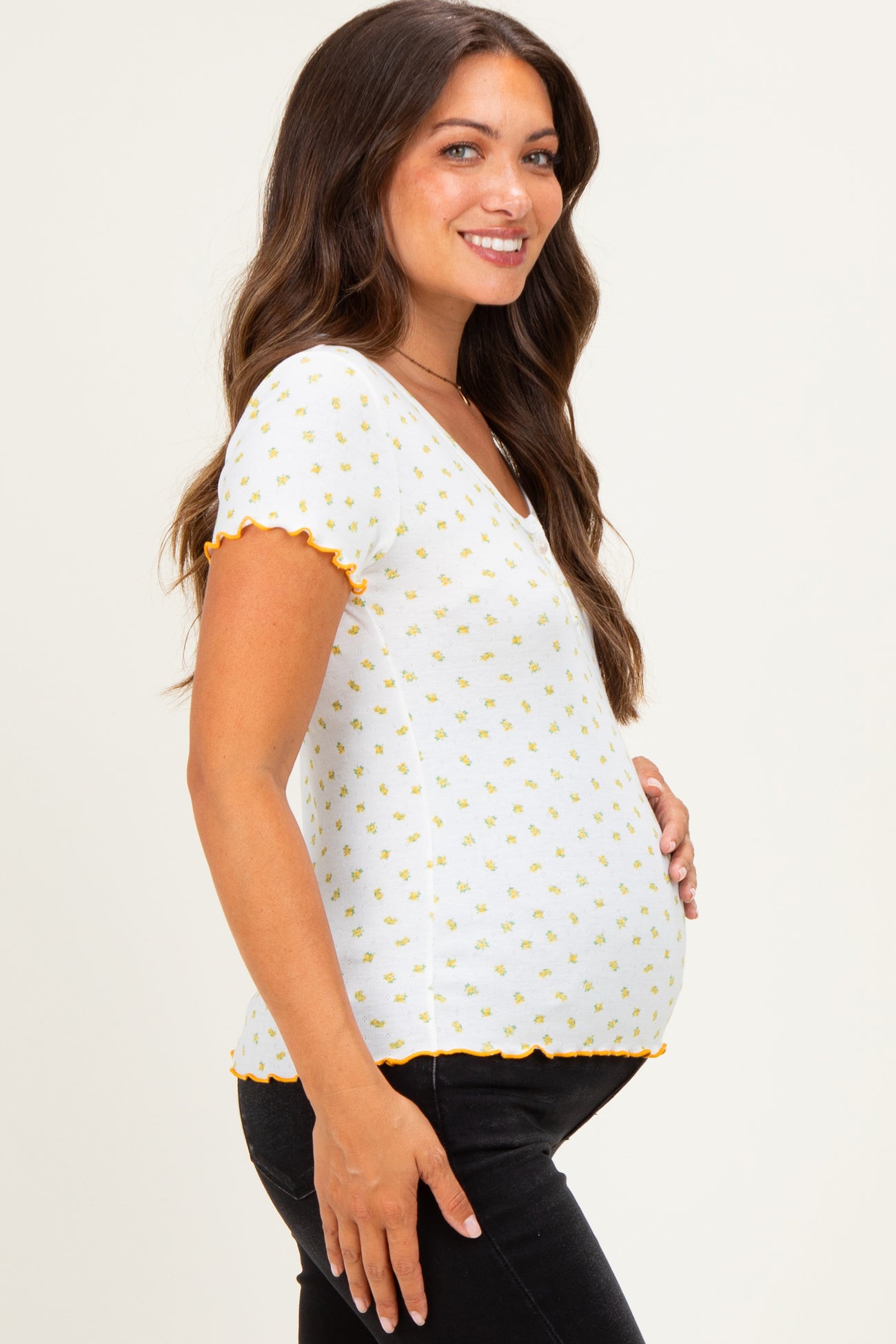 woman wearing a white short sleeve maternity top with yellow floral patterns and scalloped edges, showcasing her baby bump