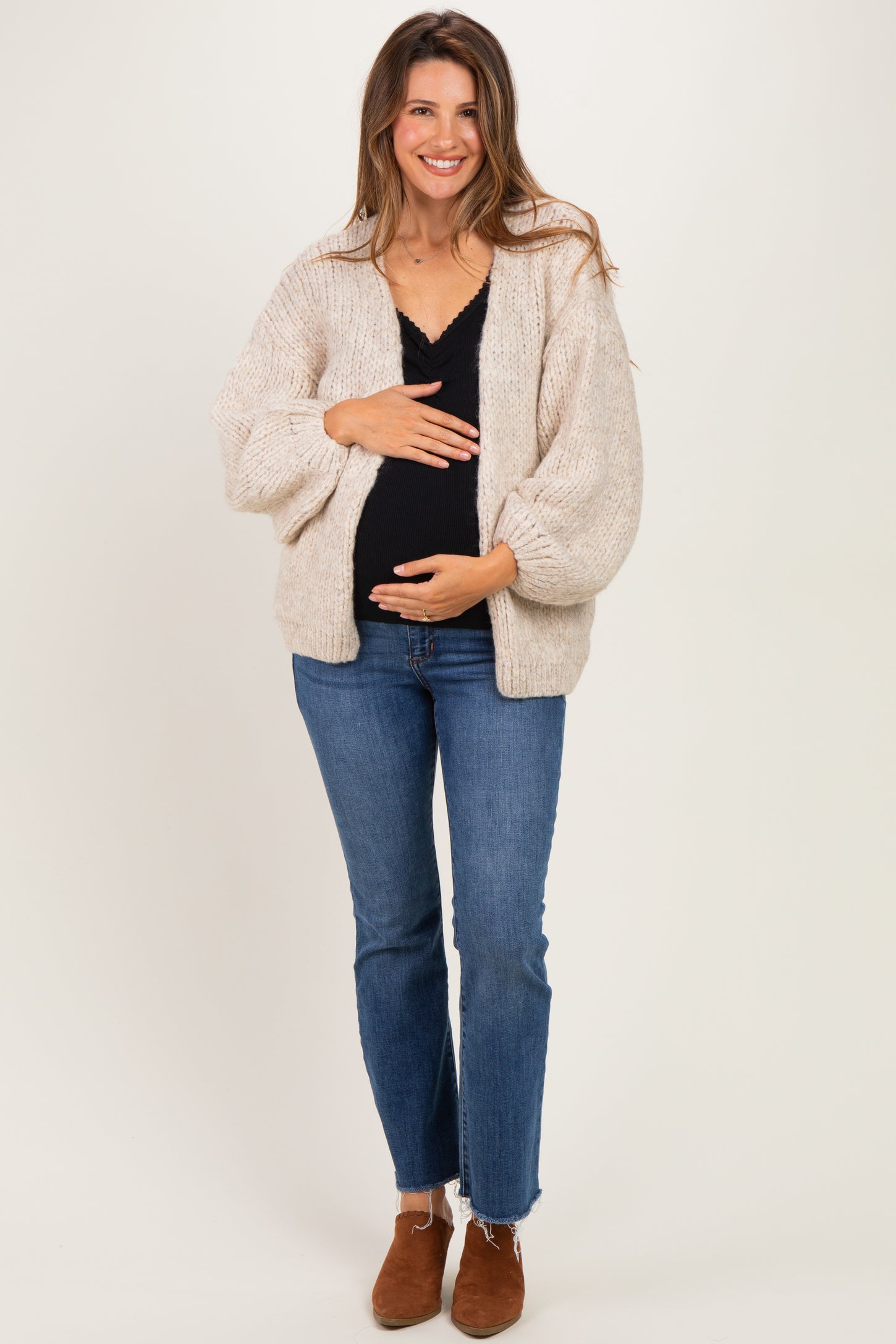 woman wearing a beige knitted maternity cardigan and black top, holding her baby bump with a smile