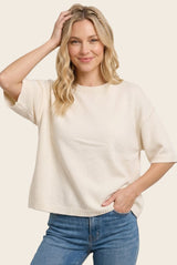 woman wearing a cream short sleeve knit sweater and blue jeans, smiling and posing with one hand in her hair