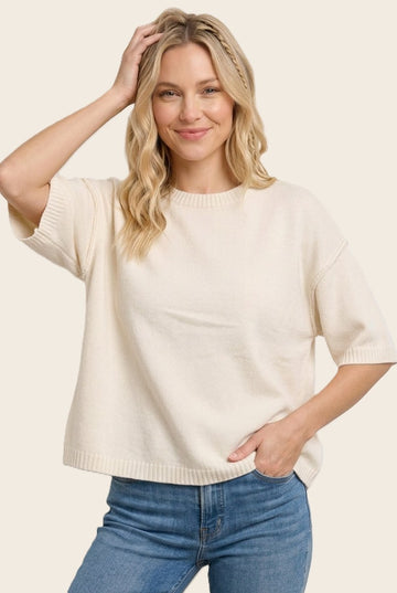 woman wearing a cream short sleeve knit sweater and blue jeans, smiling and posing with one hand in her hair