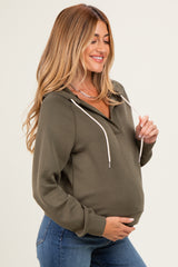 woman wearing an olive green maternity pullover hoodie with a front pocket and drawstring hood, holding her baby bump
