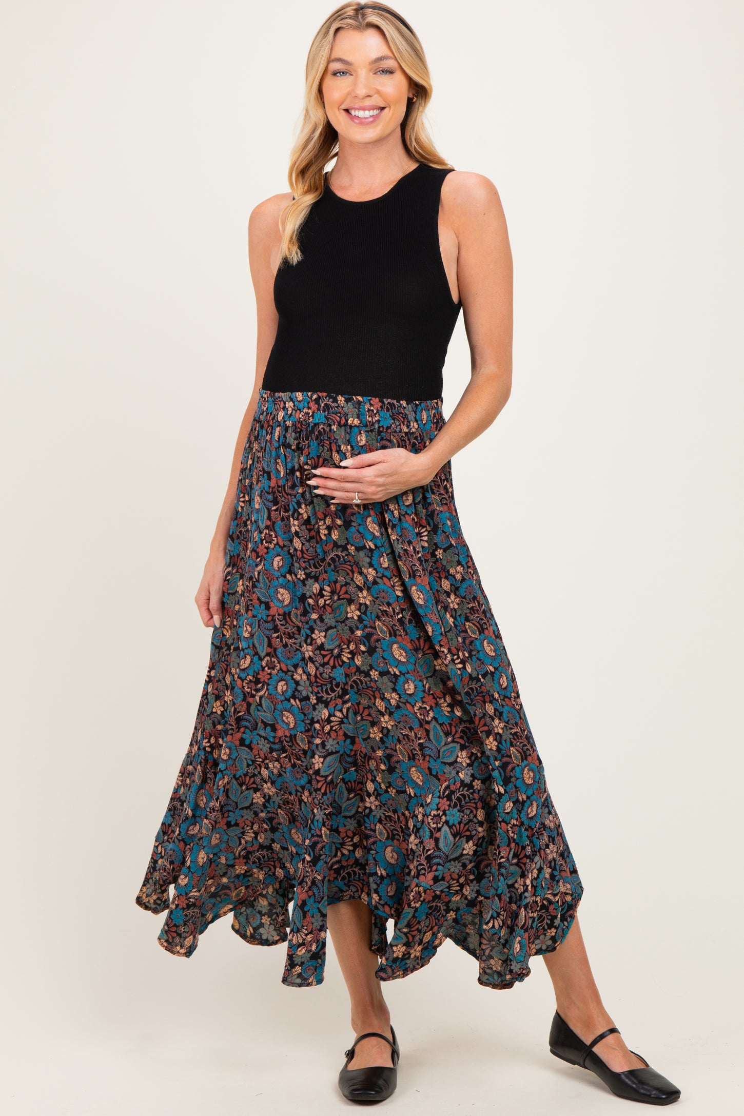 woman wearing a black sleeveless top and a floral print maternity maxi skirt, gently holding her baby bump